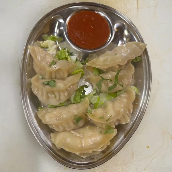 A metal plate of steamed dumplings with lettuce and cilantro, served with a side of red sauce.