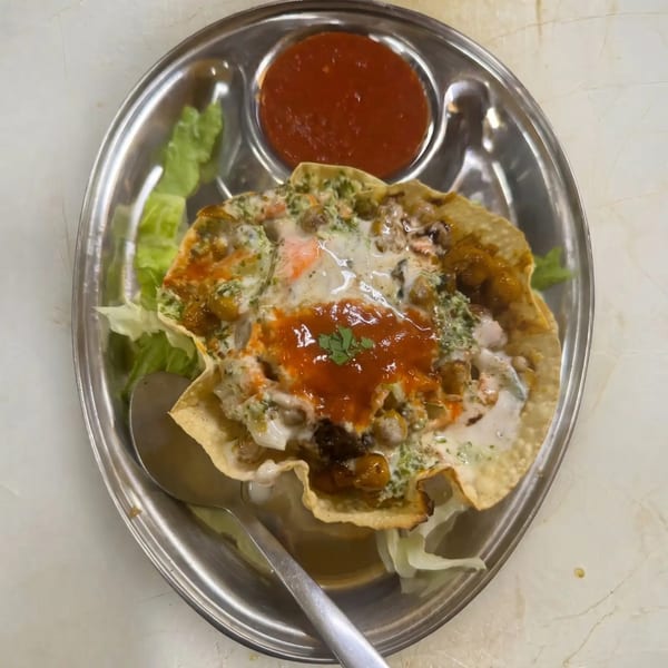 A metal plate holds a crispy tortilla bowl filled with chickpeas, sauces, and a side of red salsa.