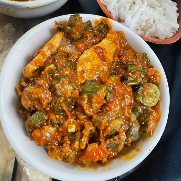 A white bowl filled with a rich, red stew containing okra, vegetables, and chicken, with white rice in the background.