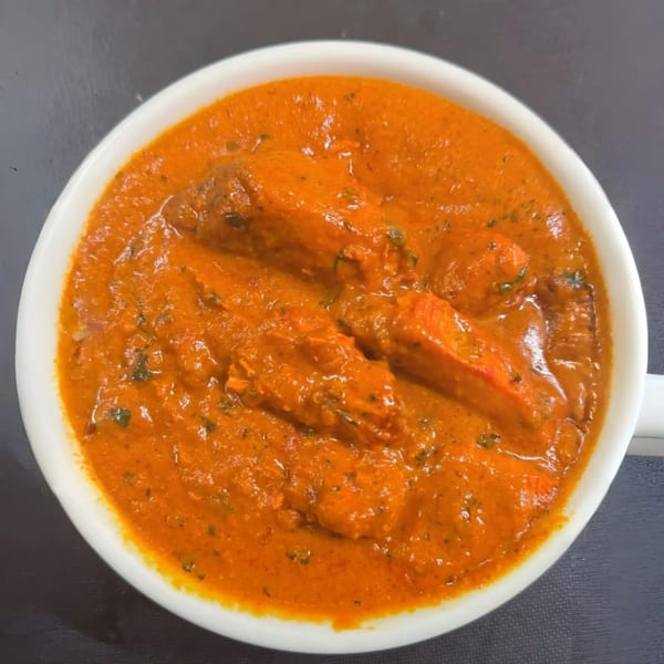 A white bowl filled with vibrant orange butter chicken, garnished with herbs, on a dark surface.